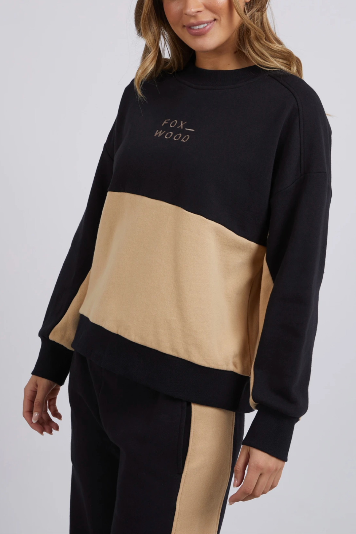 A woman wears the Foxwood Offhand Crew, a color-blocked 100% cotton fleece sweatshirt with "FOX WOOD" on the chest, paired with matching black and beige panel pants for easy lounge comfort.