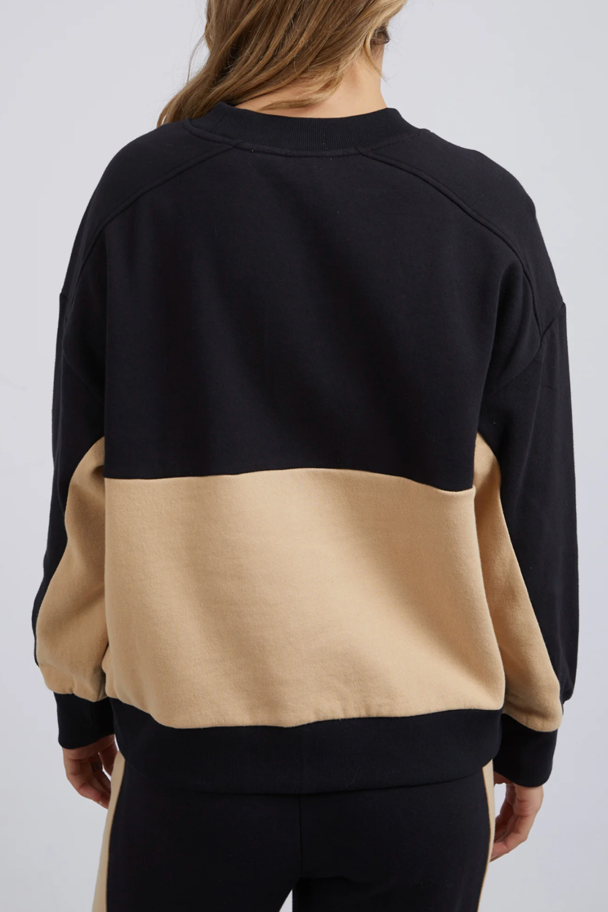 A person with long hair wears a color-blocked Foxwood Offhand Crew sweatshirt of plush cotton fleece, featuring a black upper and beige lower section. The individual faces away, highlighting the cozy back of this lounge-comfy ensemble.