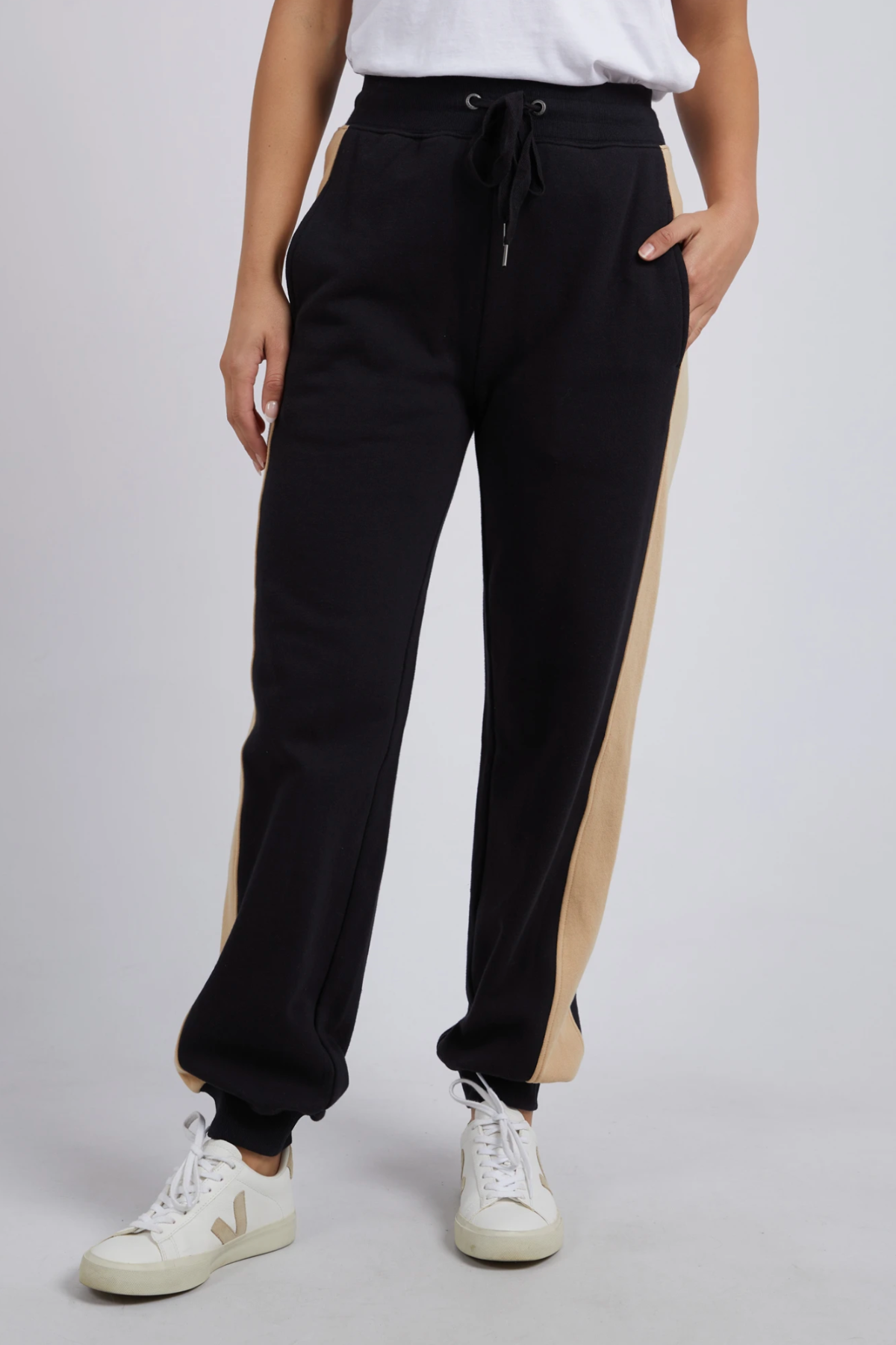 Wearing Foxwood Offhand Pant black sweatpants with tan side stripes, a white tee, and white sneakers, a person stands with one hand in their pocket, showcasing 100% cotton fleece comfort against a plain background.