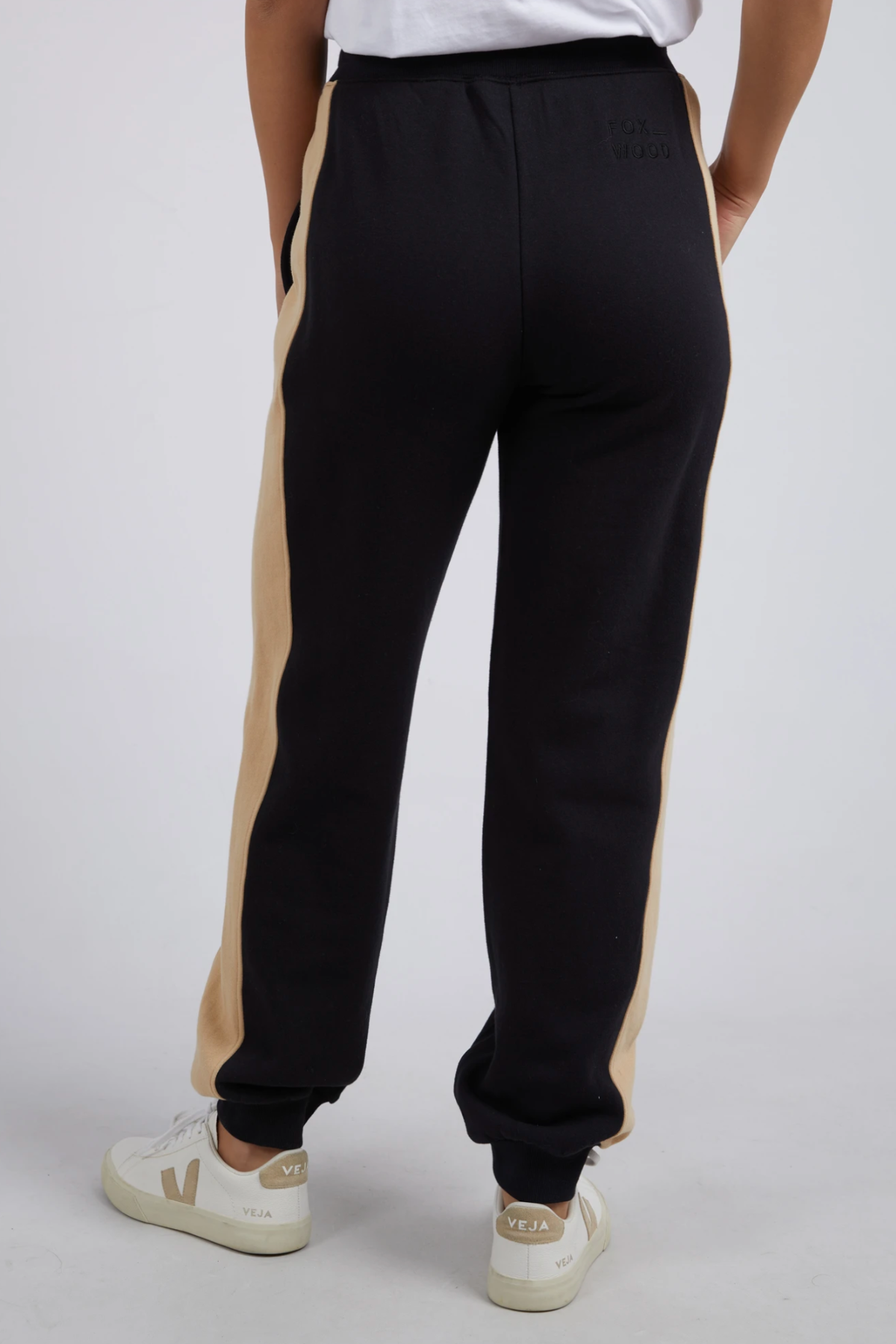 Person wearing black Foxwood Offhand Pant with beige side stripes, a white 100% cotton fleece T-shirt, and white Veja sneakers stands against a light background. The back view highlights the lounge comfort of the Offhand Pant.