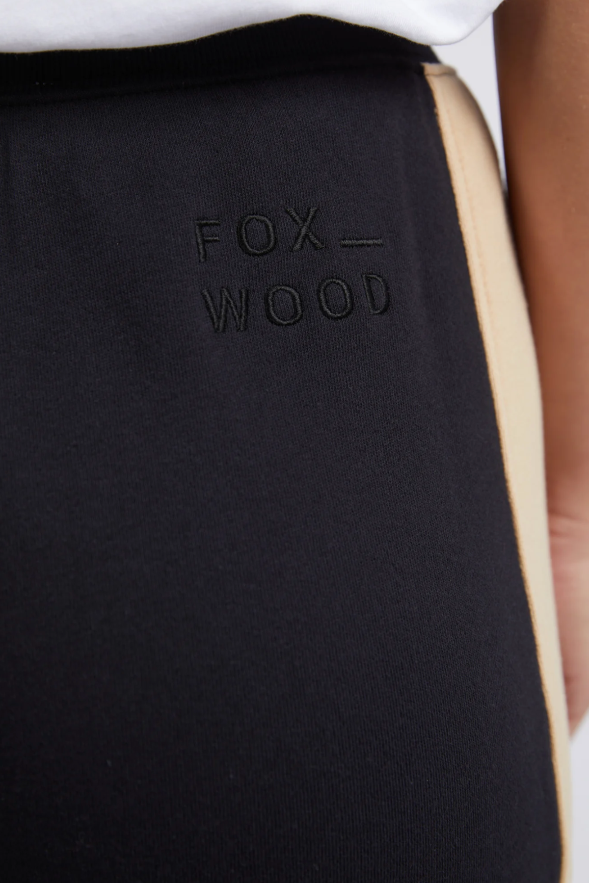 The Foxwood Offhand Pant features a beige side stripe, "FOX WOOD" embroidery on the hip, and a relaxed fit cotton fleece design. Shown paired with a white shirt.