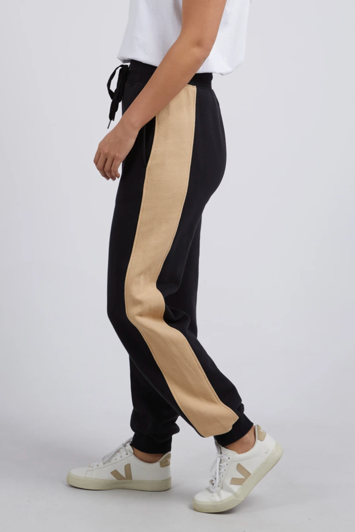 Wearing the Foxwood Offhand Pant—black joggers featuring a beige side stripe and made from 100% cotton fleece—a person pairs them with a white T-shirt and white sneakers, standing sideways against a plain light background.