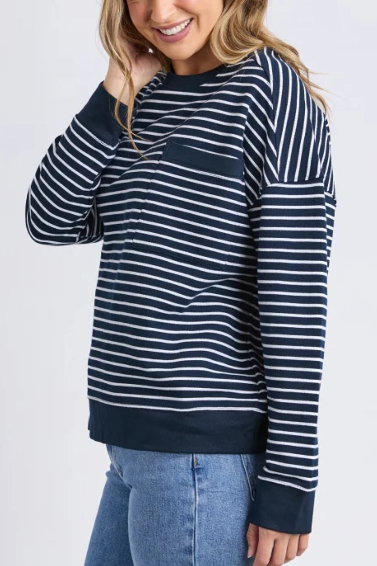 A woman smiles, touching her hair, wearing the Foxwood Original Stripe Crew in navy blue and white with a relaxed fit, paired with light blue jeans against a plain light background.
