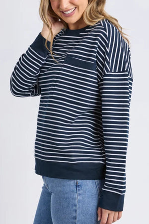 A woman smiles, touching her hair, wearing the Foxwood Original Stripe Crew in navy blue and white with a relaxed fit, paired with light blue jeans against a plain light background.