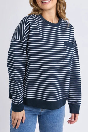 A woman wears the Foxwood Original Stripe Crew, a navy and white relaxed fit jumper with a chest pocket, paired with blue jeans. She smiles against a neutral background—perfect for effortless winter style.