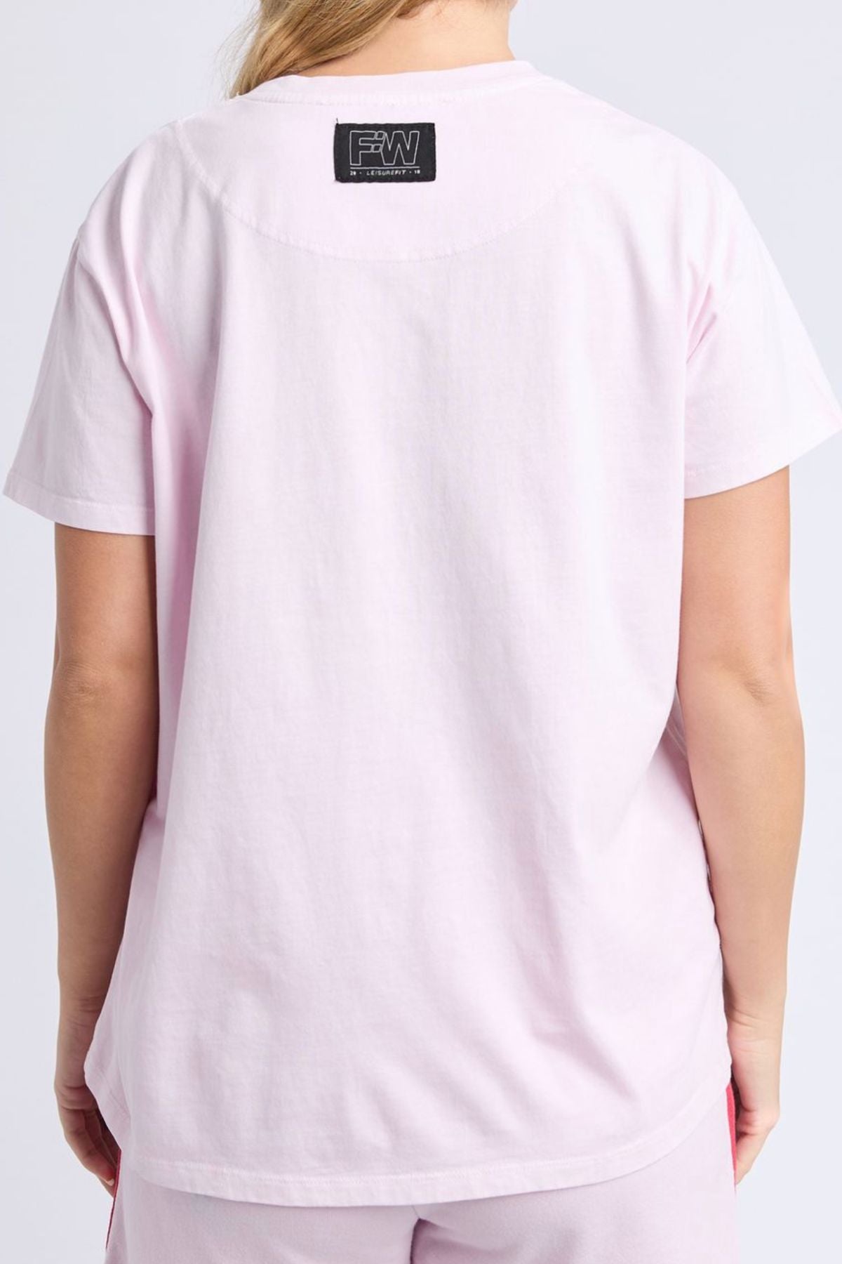 A person is seen from the back wearing a light pink Foxwood Palermo Tee and matching pants. The relaxed-fit tee features a black rectangular label with white text near the neckline.