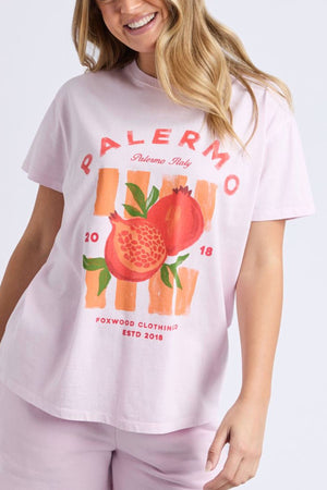 A woman smiles in a relaxed-fit Foxwood Palermo Tee—a light pink t-shirt with pomegranates, orange stripes, and “PALERMO Italy 2018” from Foxwood Clothing Co.