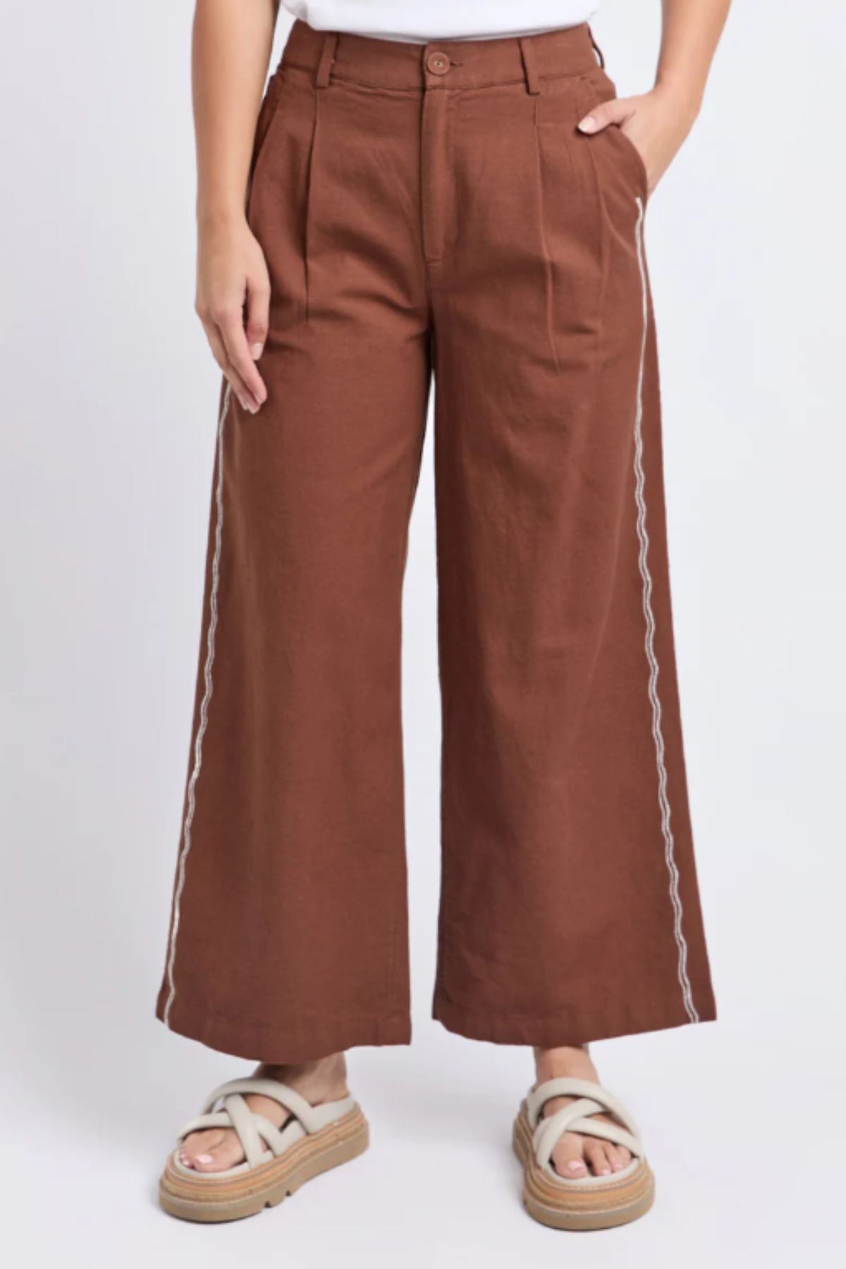 Someone stands against a plain light background wearing the Foxwood Paradise Pant—high-waisted, relaxed-fit trousers with white side stitching—paired with a white top and beige platform sandals.