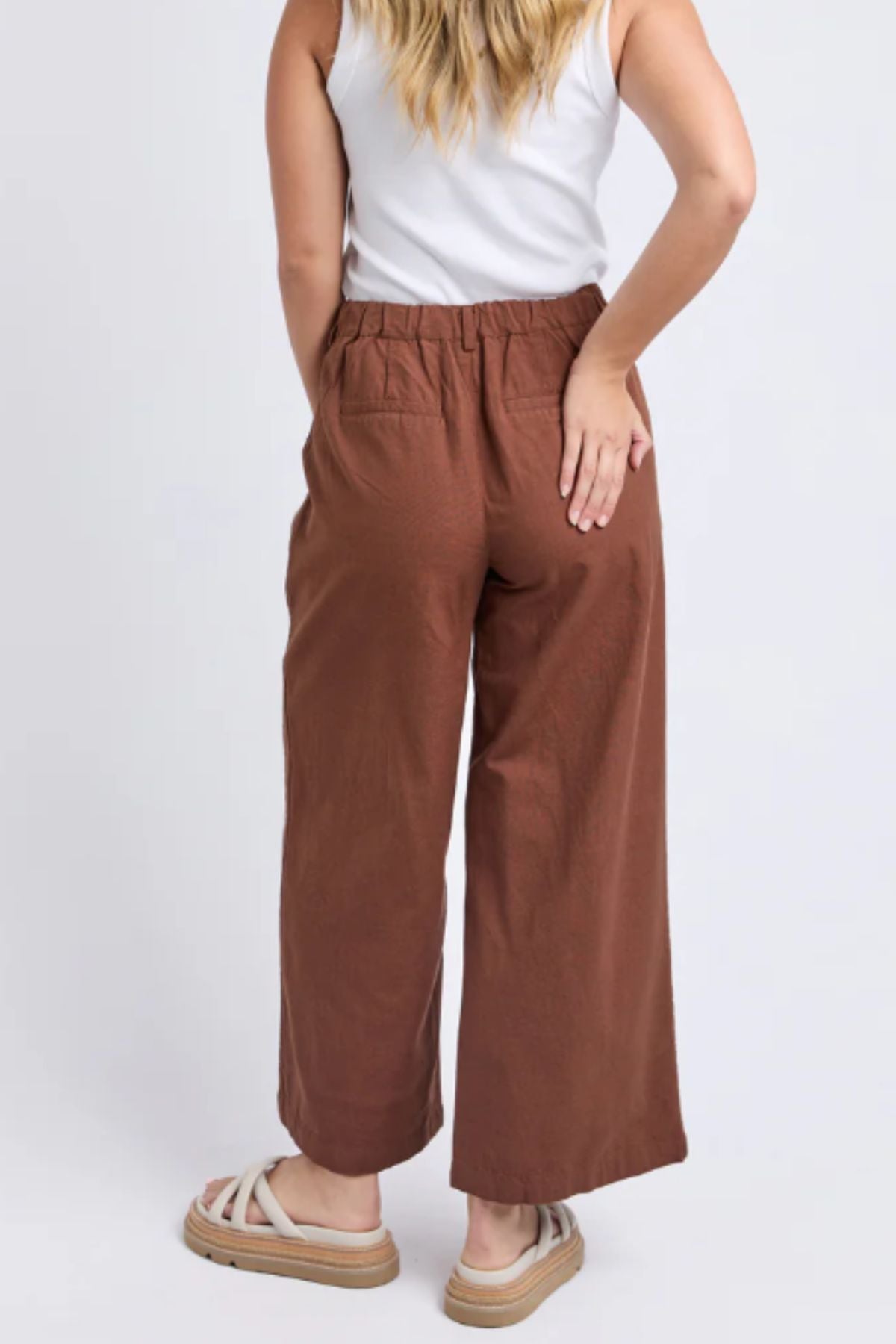 A woman faces away, wearing Foxwood Paradise Pant—relaxed-fit with elastic waist—paired with a white sleeveless top and beige sandals. Her left hand rests on her hip, and long wavy hair cascades over her shoulders.