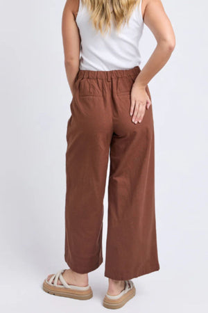 A woman faces away, wearing Foxwood Paradise Pant—relaxed-fit with elastic waist—paired with a white sleeveless top and beige sandals. Her left hand rests on her hip, and long wavy hair cascades over her shoulders.