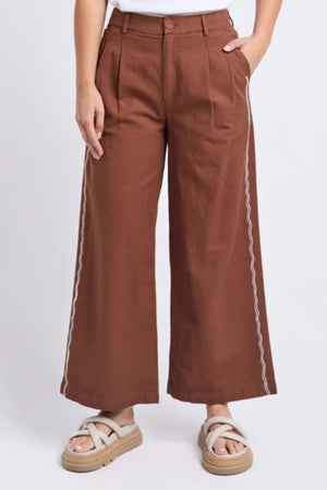 Someone stands against a plain light background wearing the Foxwood Paradise Pant—high-waisted, relaxed-fit trousers with white side stitching—paired with a white top and beige platform sandals.