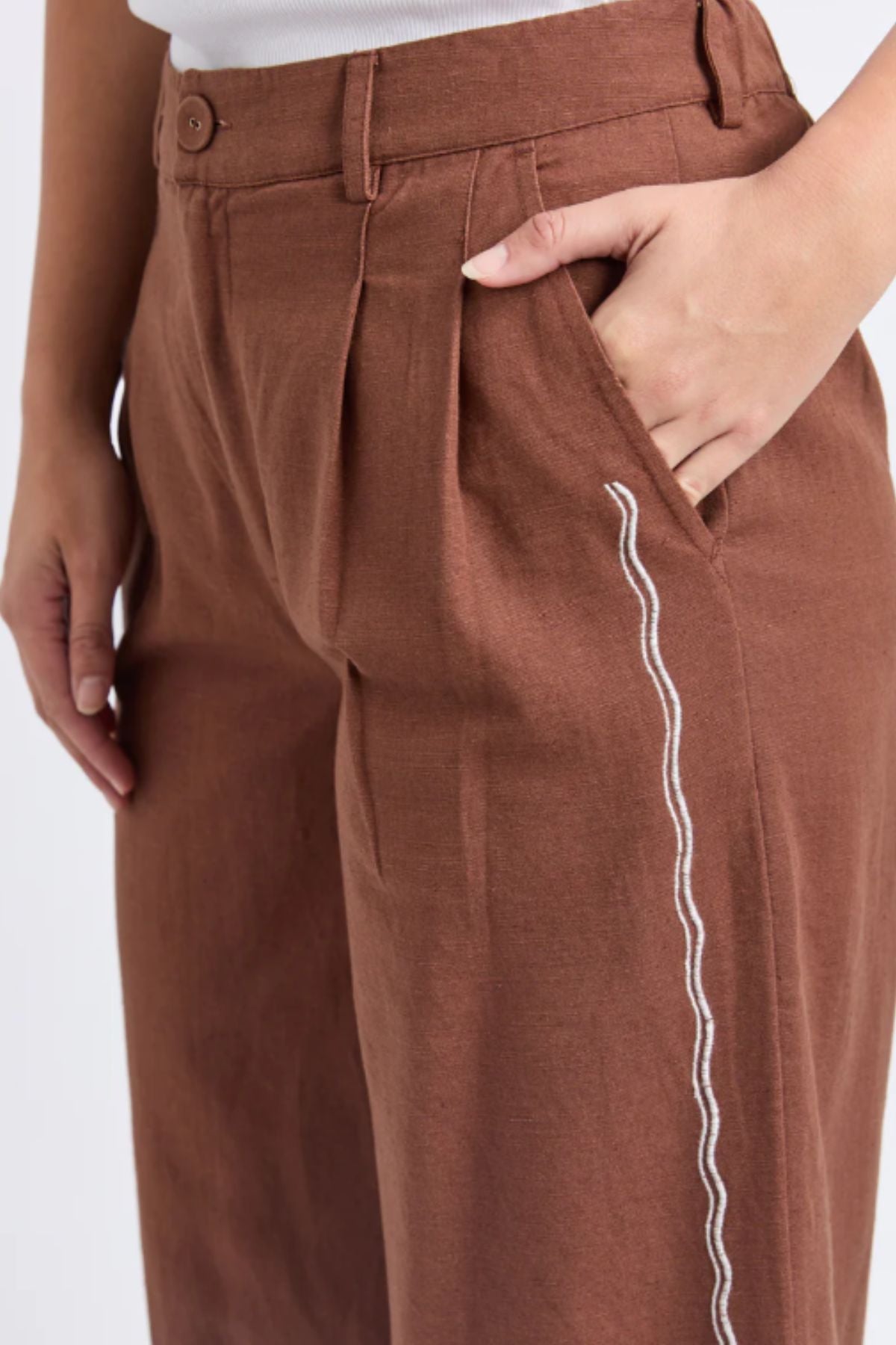 A person wears the Foxwood Paradise Pant in brown, featuring a white wavy line along the side pocket, paired with a tucked-in white top. One hand is casually placed inside the pocket.