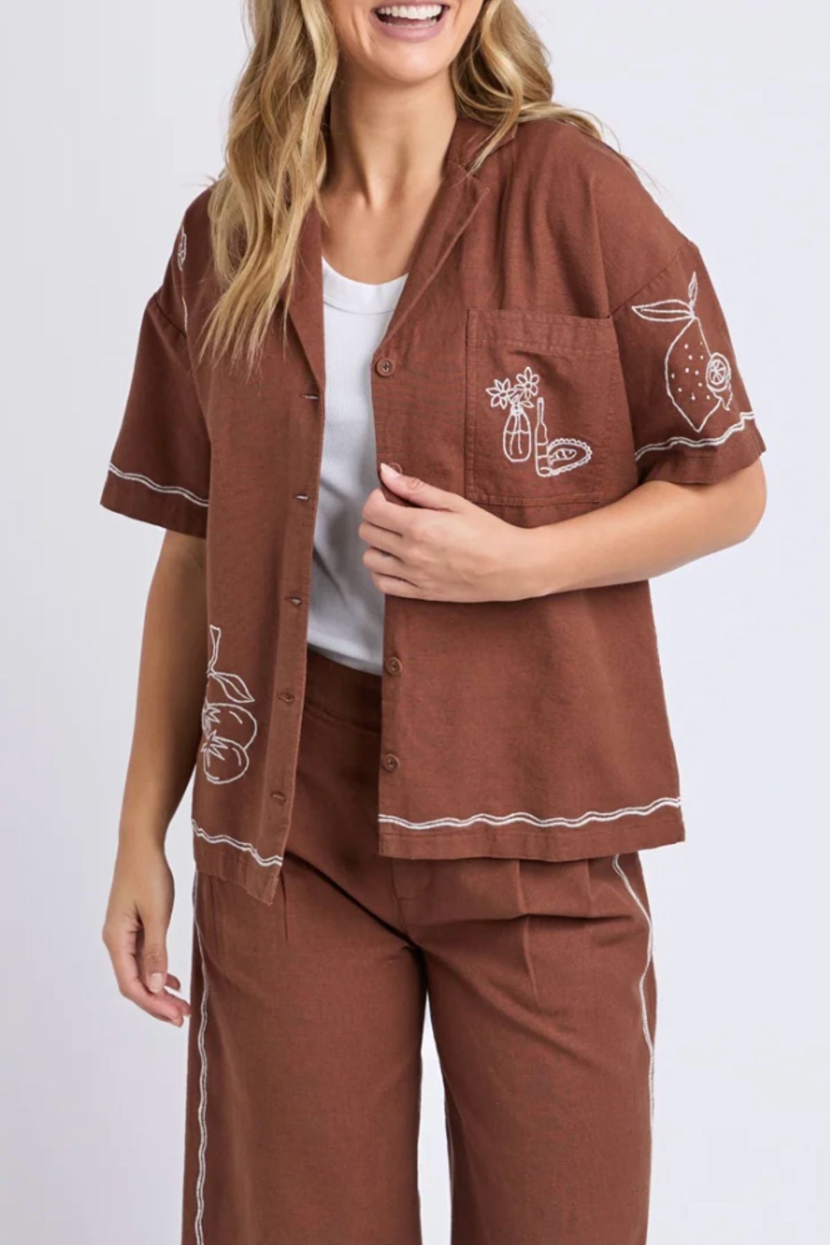 A person with wavy, shoulder-length hair smiles while wearing the Foxwood Paradise Shirt—a relaxed-fit, brown short-sleeve embroidered top with white stitched fruit and plant designs—layered over a white top and paired with matching pants.