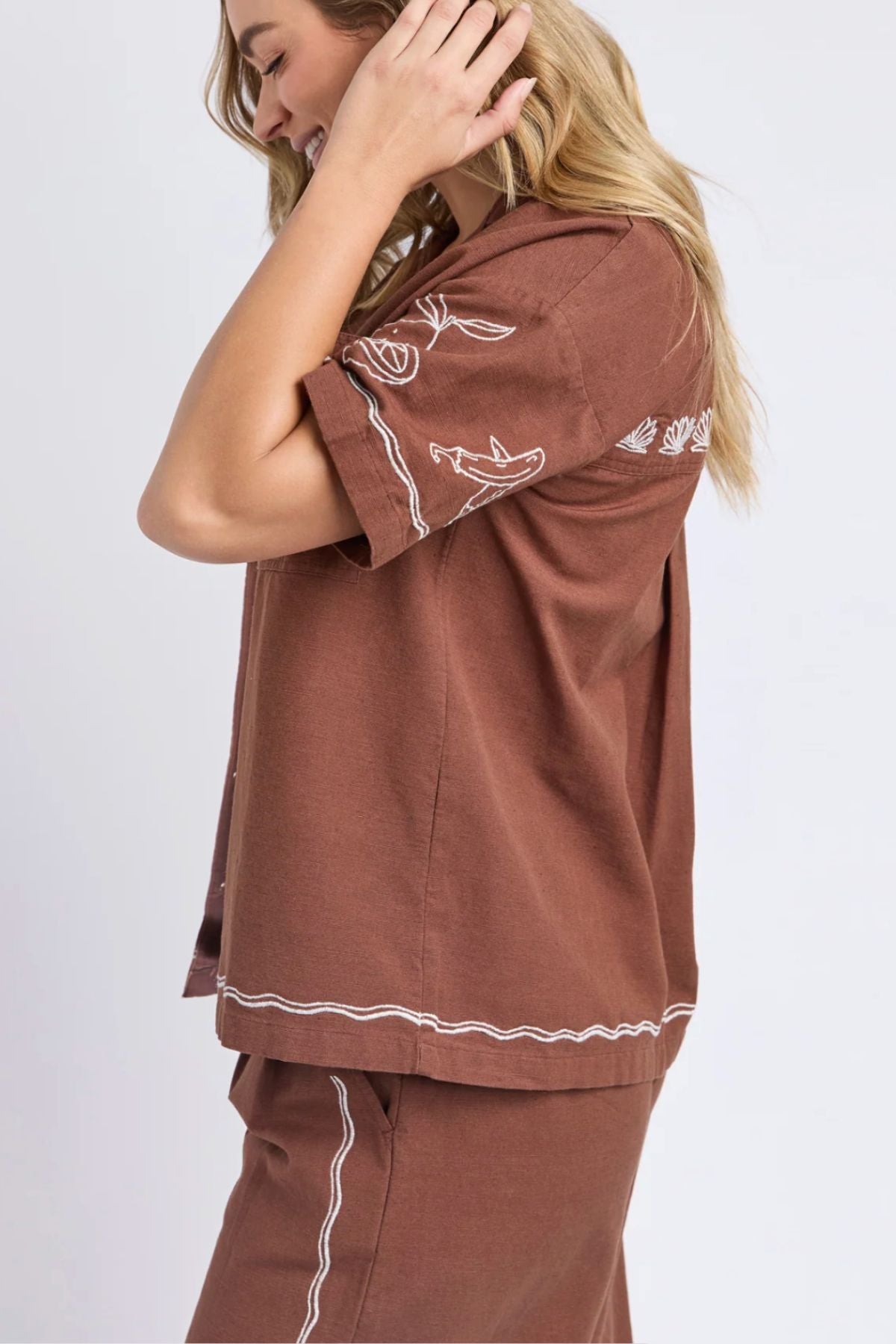 A woman smiles and touches her hair while wearing the Foxwood Paradise Shirt with matching shorts, both featuring white stitched designs on the sleeves, hem, and shorts.