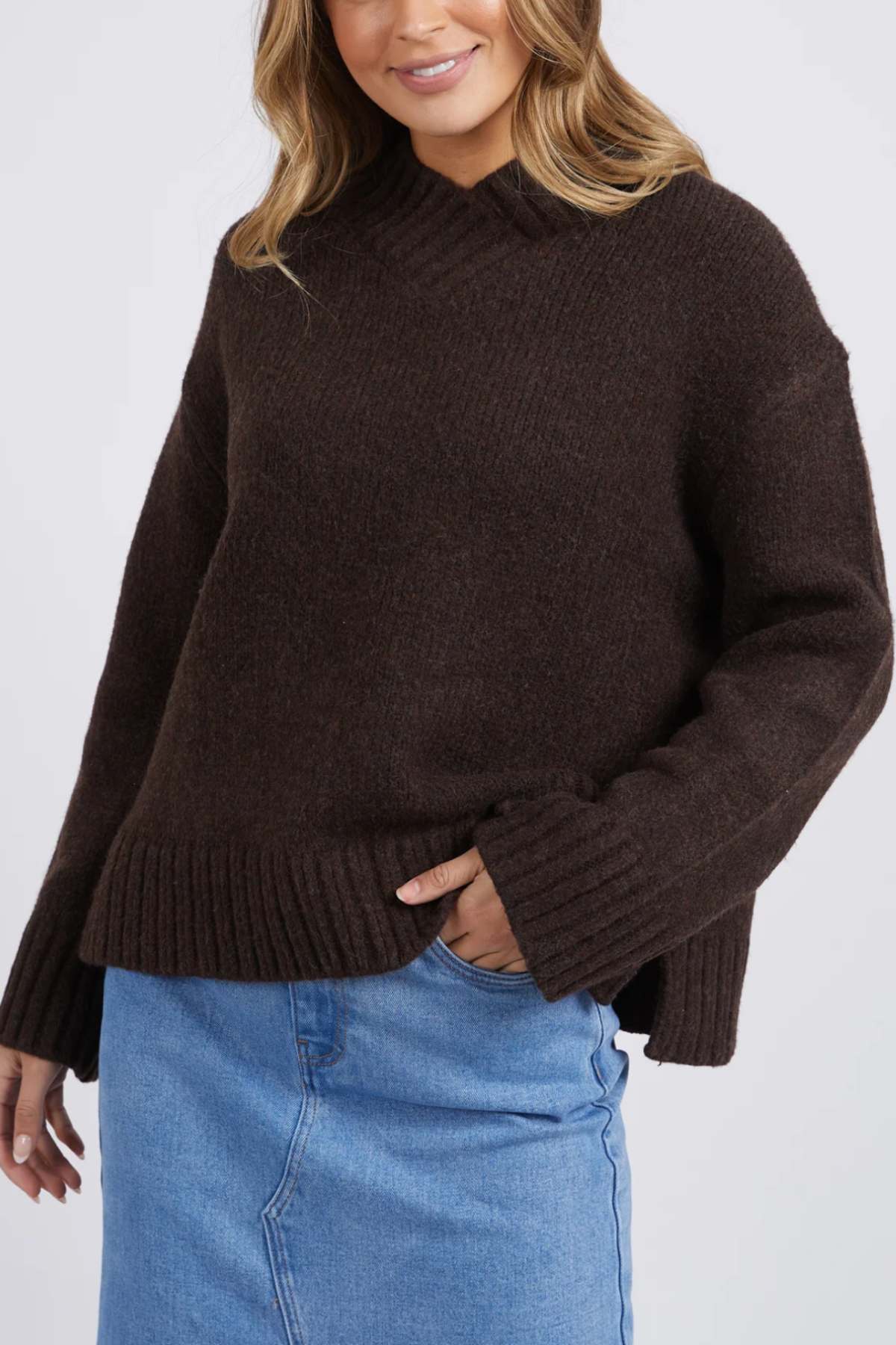 A woman smiles in the Foxwood Pepper Knit, a dark brown long-sleeve sweater with a ribbed hem and cozy blend, paired with a light blue denim skirt against a plain light background.