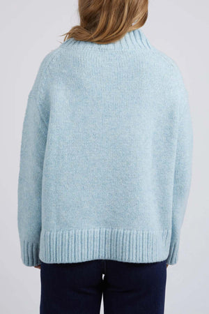 A person with light brown hair is seen from the back wearing the Foxwood Pepper Knit, a cozy blend light blue sweater featuring a vee neckline, relaxed fit, and ribbed hem. Paired with dark pants against a plain light-colored background.