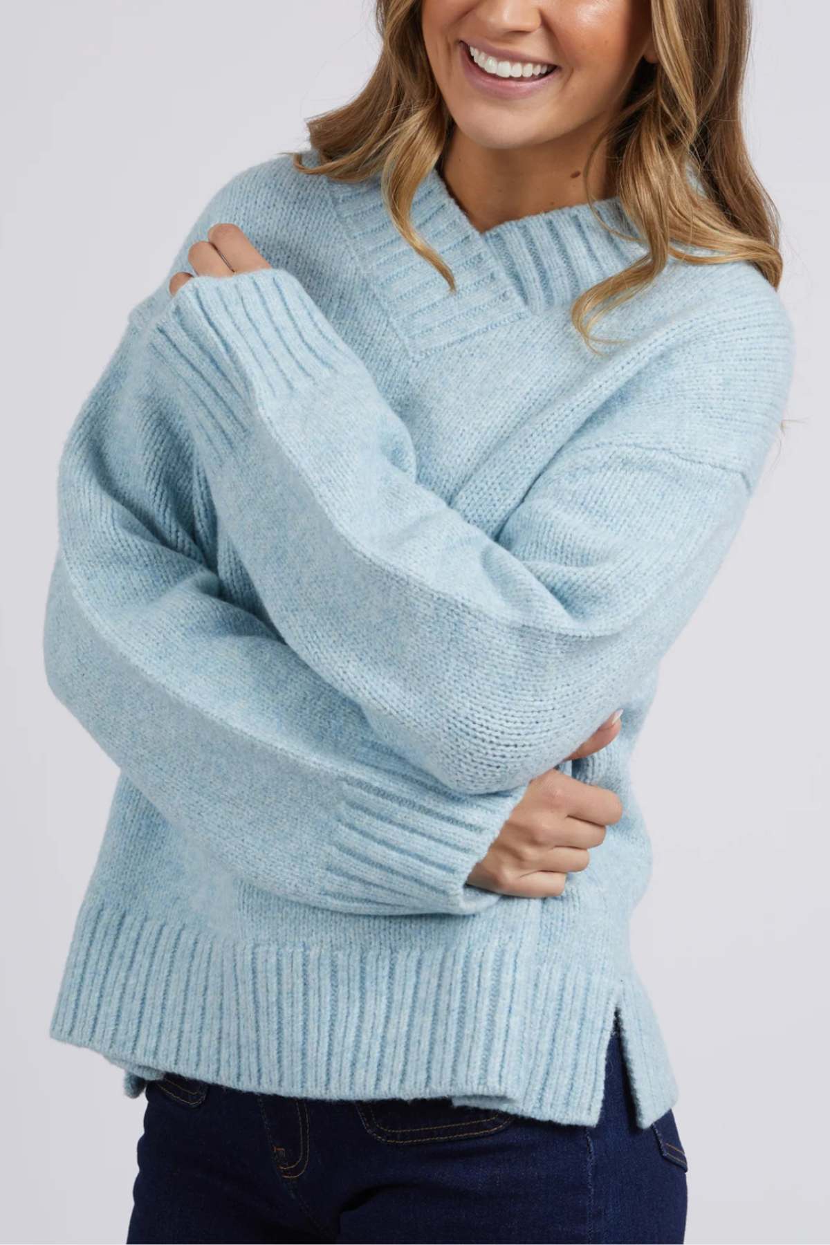 Wrapped in the oversized Foxwood Pepper Knit, a person exudes comfort and style. The ribbed sweater's light blue hue and relaxed fit are complemented by a chic Vee neckline. Their long hair gracefully falls over folded arms against a plain, light background that enhances their tranquil demeanor.