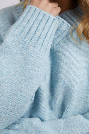 A person is wearing the Foxwood Pepper Knit, a light blue chunky knit sweater with ribbed cuffs and a thick collar. The cozy blend has a relaxed fit, with long hair resting on their chin, partially covered by the sleeve.
