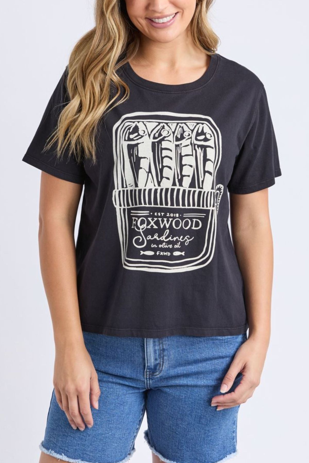 Wearing the Foxwood Pesce Tee, a coastal-inspired black cotton T-shirt with a sardine can graphic labeled "Foxwood Sardines," and blue denim shorts, a person stands against a plain background.