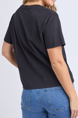 A woman models the Foxwood Pesce Tee, a black short-sleeve t-shirt, with blue jeans. She is pictured from the back, head turned slightly to the side, against a simple light background.
