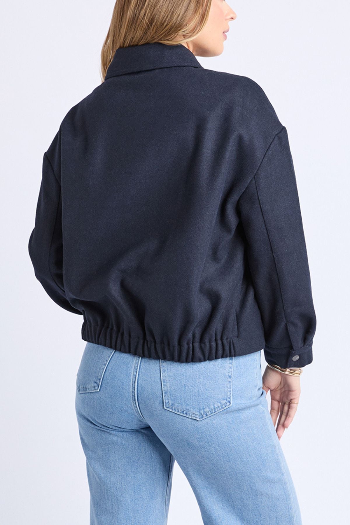 A woman with light brown hair wears the Foxwood Pippa Jacket, a dark navy wool blend with an elastic waistband, paired with light blue high-waisted jeans against a plain white background—an ideal cosy outerwear choice.