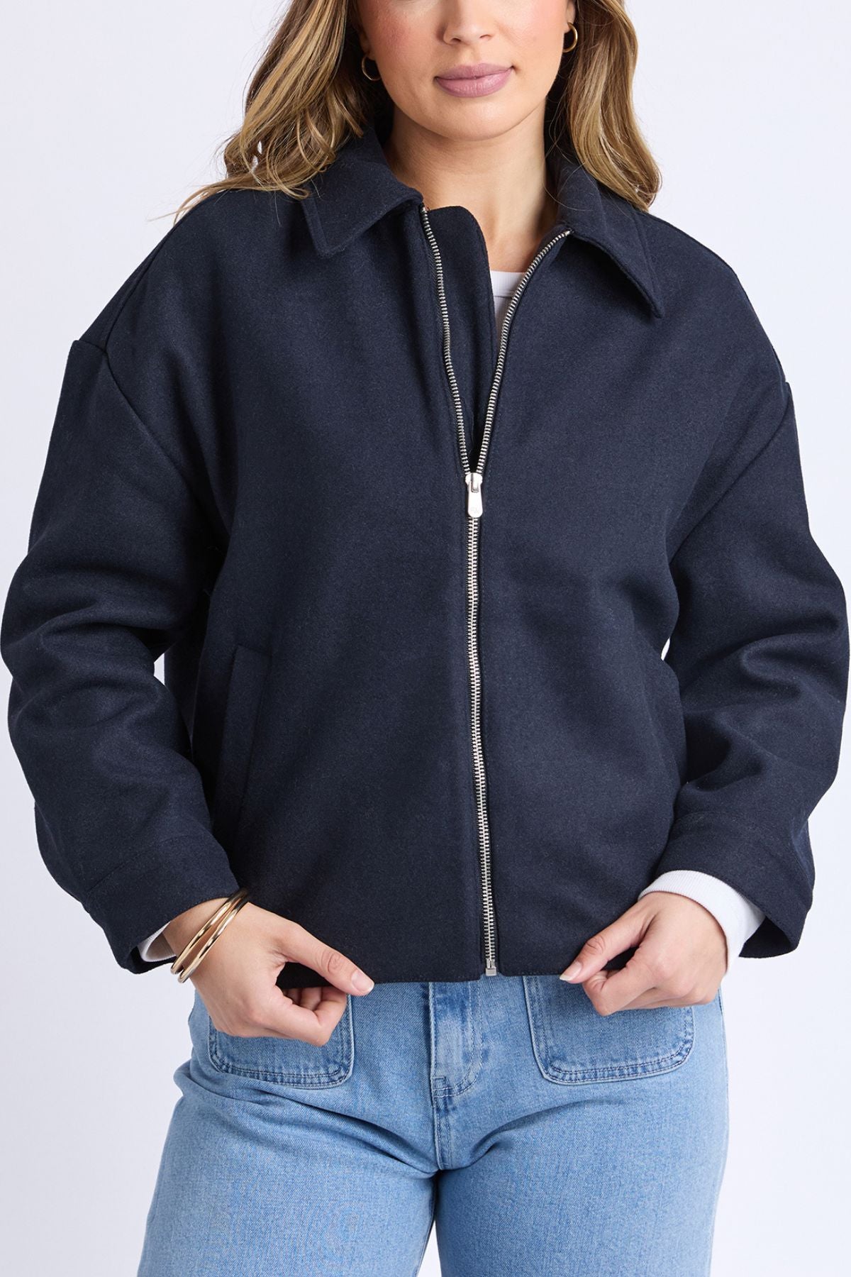 A person wearing the Foxwood Pippa Jacket—a navy blue wool blend—over a white shirt and light blue jeans. With wavy hair, gold hoop earrings, and bracelets, this cosy outerwear layer is perfect for women.