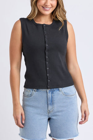 A woman smiles in comfort, wearing the Foxwood Raf Button Through Tank—sleeveless and black—paired with light blue high-waisted denim shorts with frayed hems, standing against a plain light background.