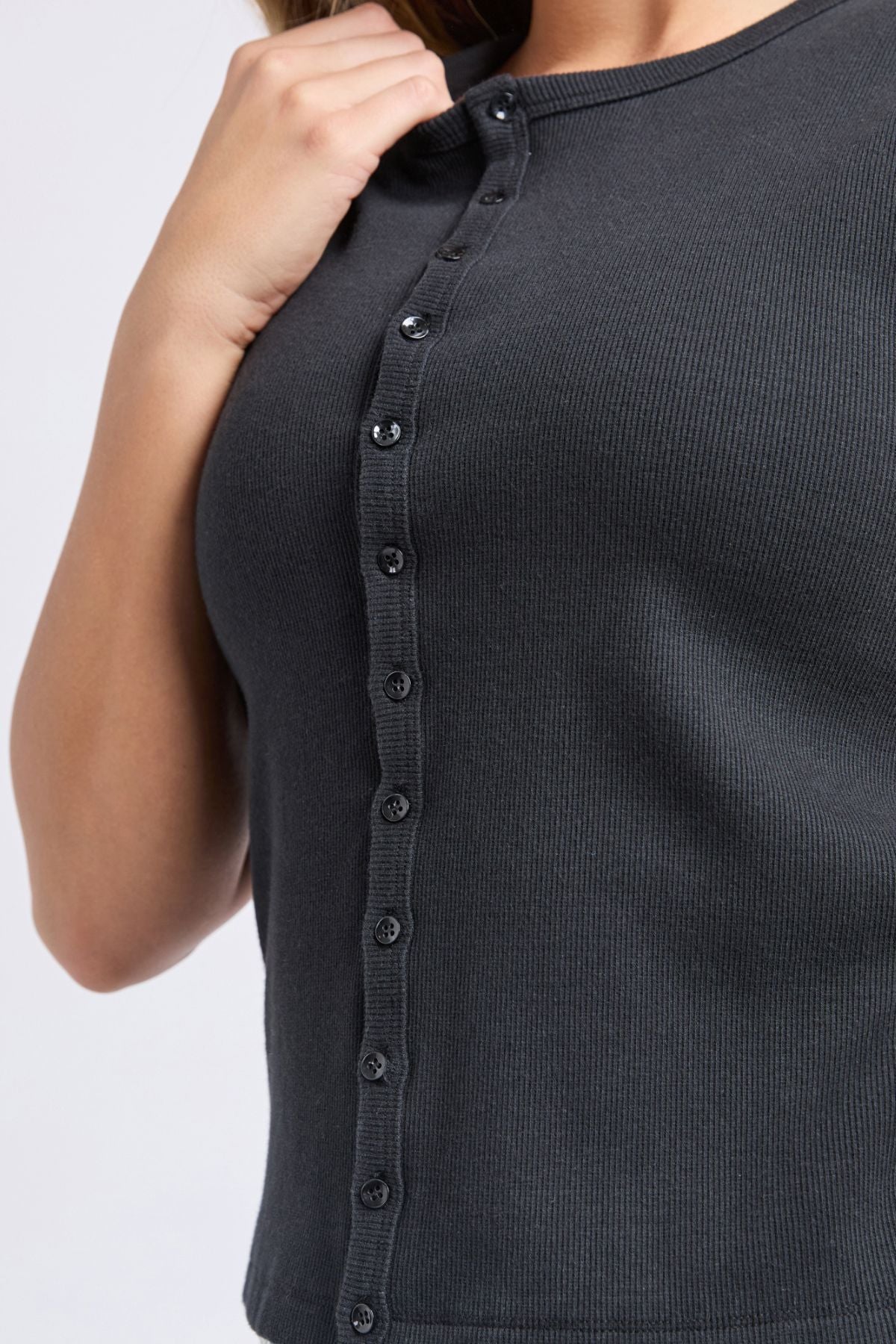 A close-up shows a person wearing the Foxwood Raf Button Through Tank in dark rib fabric, featuring short sleeves and buttons. Their raised hand near the collar highlights the tank's comfortable fit.