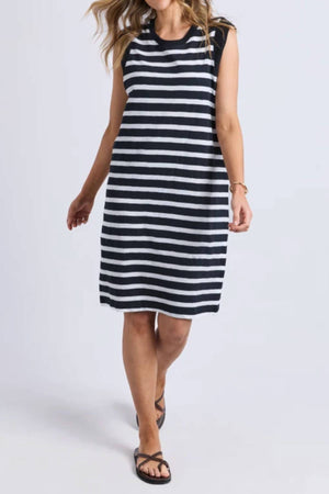A woman in a sleeveless, knee-length Foxwood Reform Stripe Dress—black and white cotton with a relaxed fit—paired with black sandals stands against a plain light background.