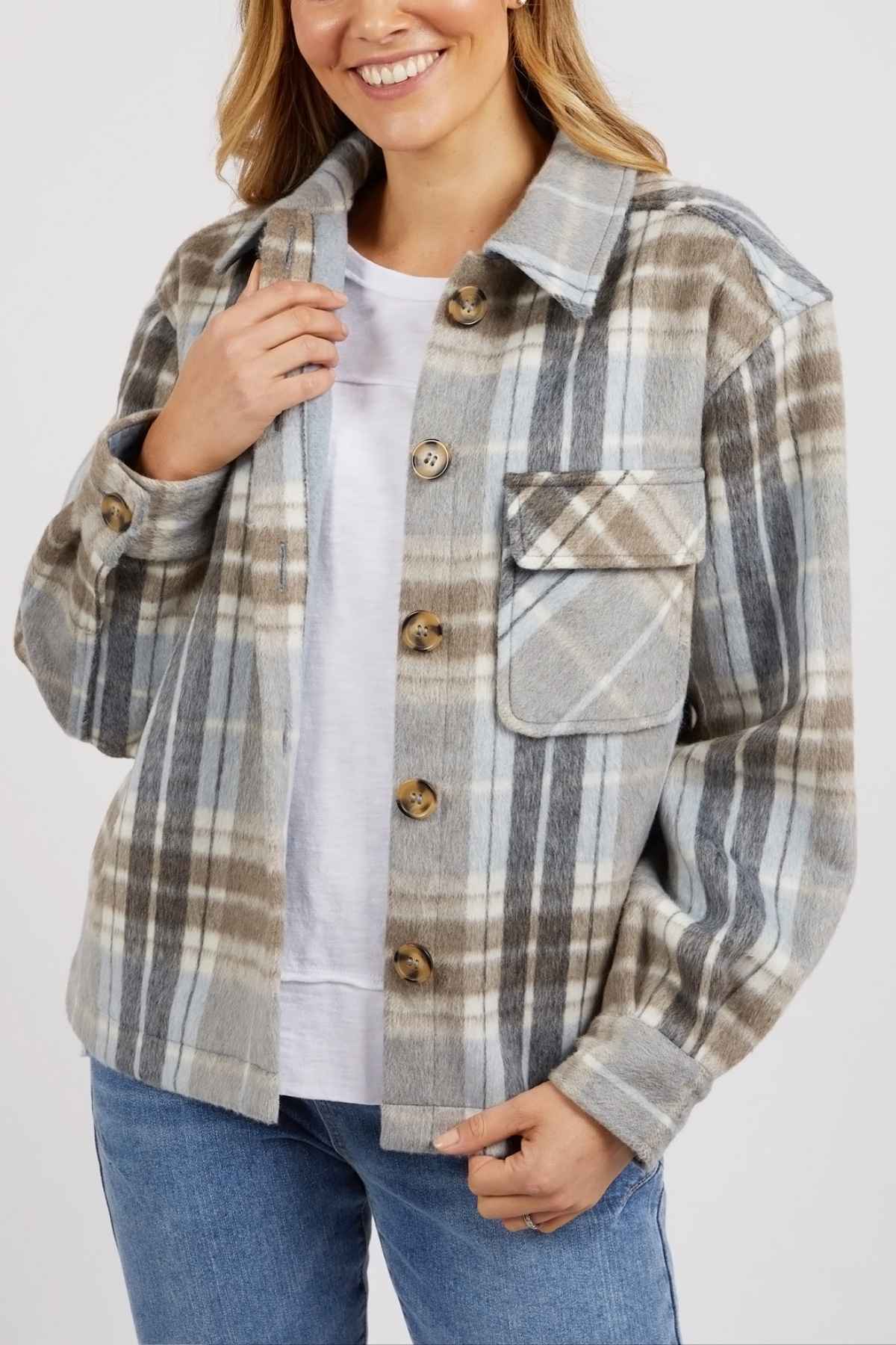 A woman smiles in the Foxwood Rhodes Check Coat—a gray, beige, and white yarn-dyed check design—styled over a white tee and blue jeans, with one hand on the collar and the other at the hem.