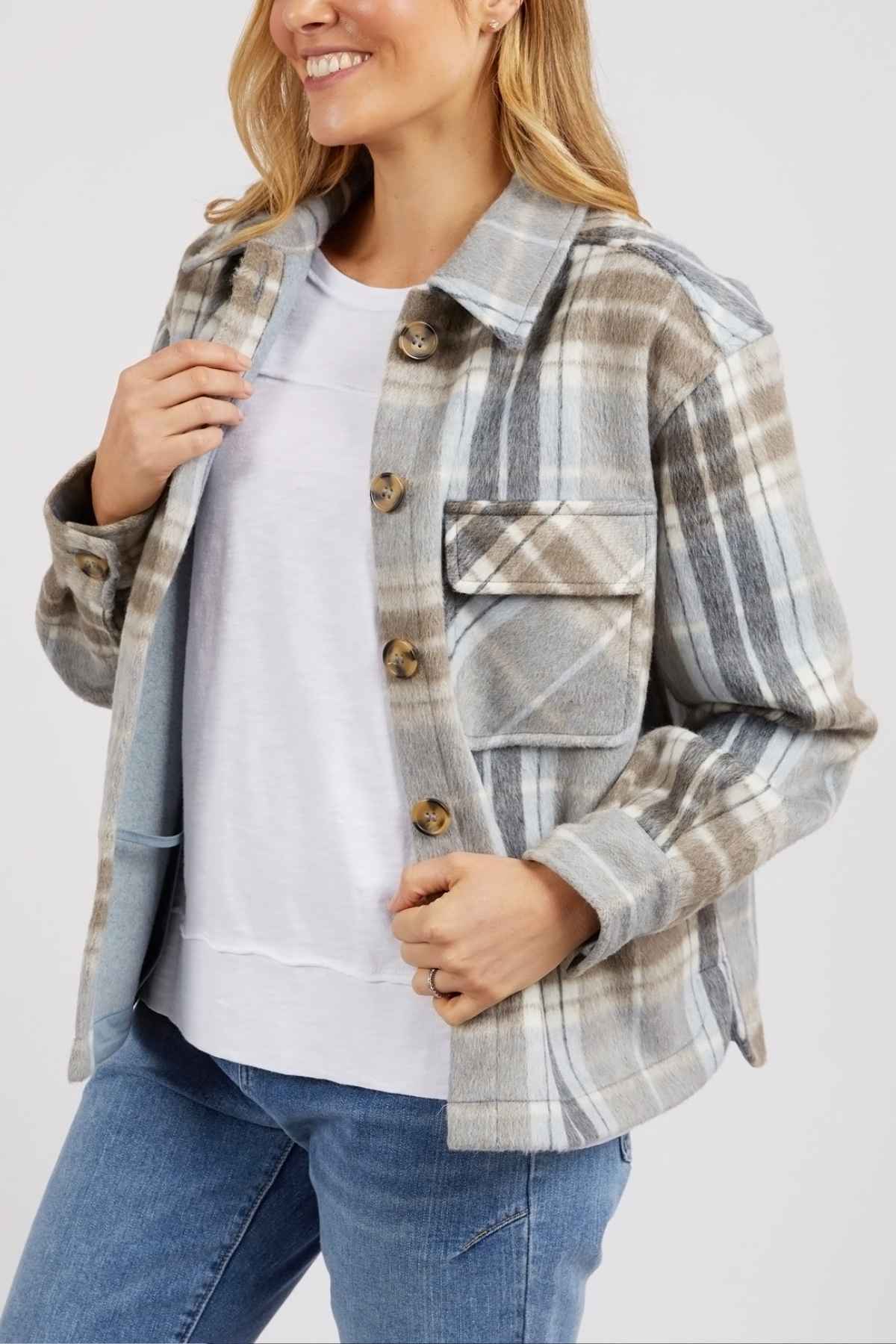 A woman smiles while holding the lapel of the Foxwood Rhodes Check Coat, styled with a white t-shirt and blue jeans. The coat features a yarn-dyed check pattern.