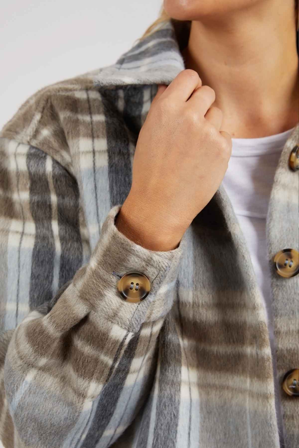 A person in the Foxwood Rhodes Check Coat, with a cozy gray and beige check wool blend, over a white top. They hold the collar with one hand while their face is partially out of frame.