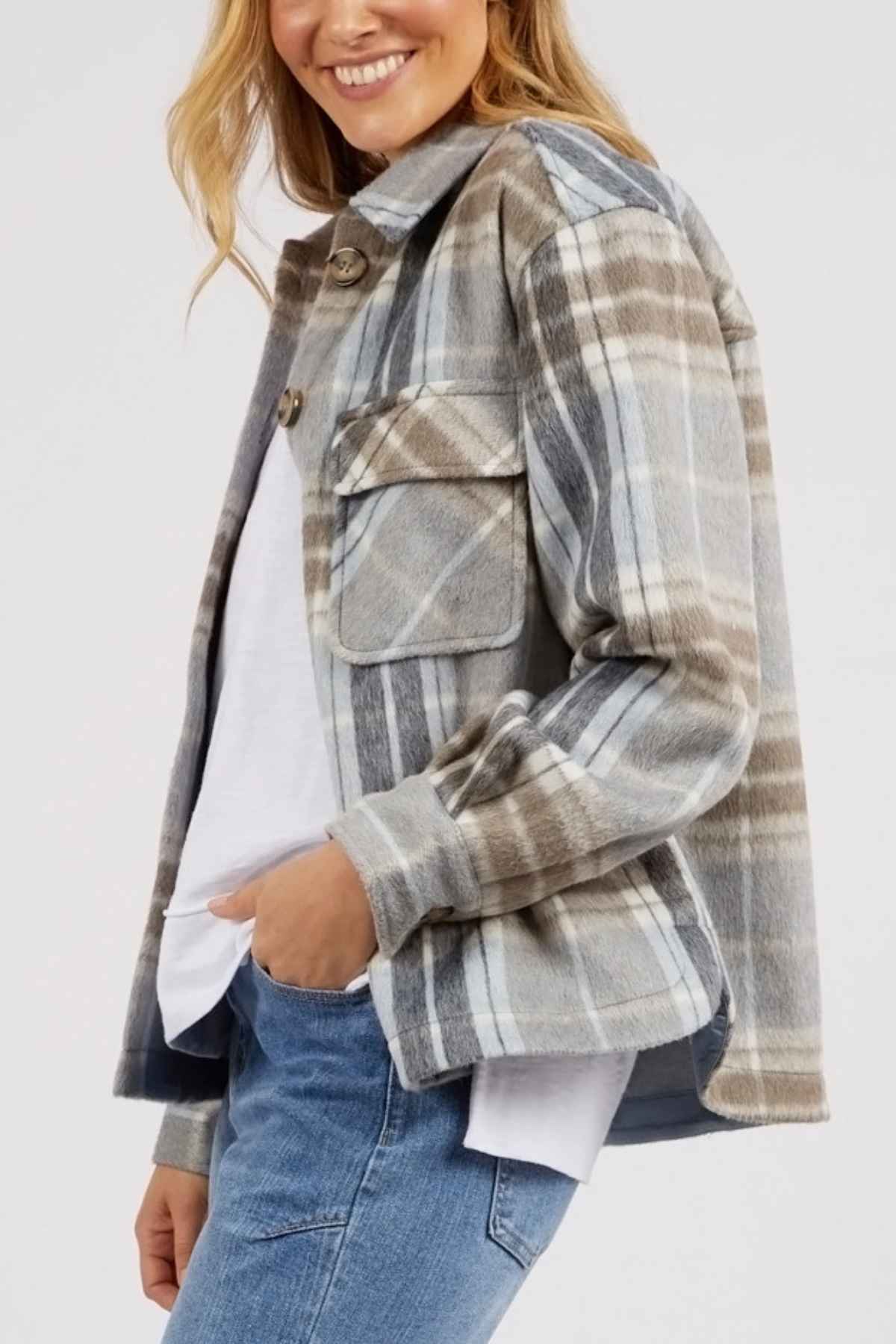 A woman smiles with one hand in her pocket, wearing the Foxwood Rhodes Check Coat—a yarn-dyed check design in gray, beige, and white—over a white shirt and blue jeans against a plain background.