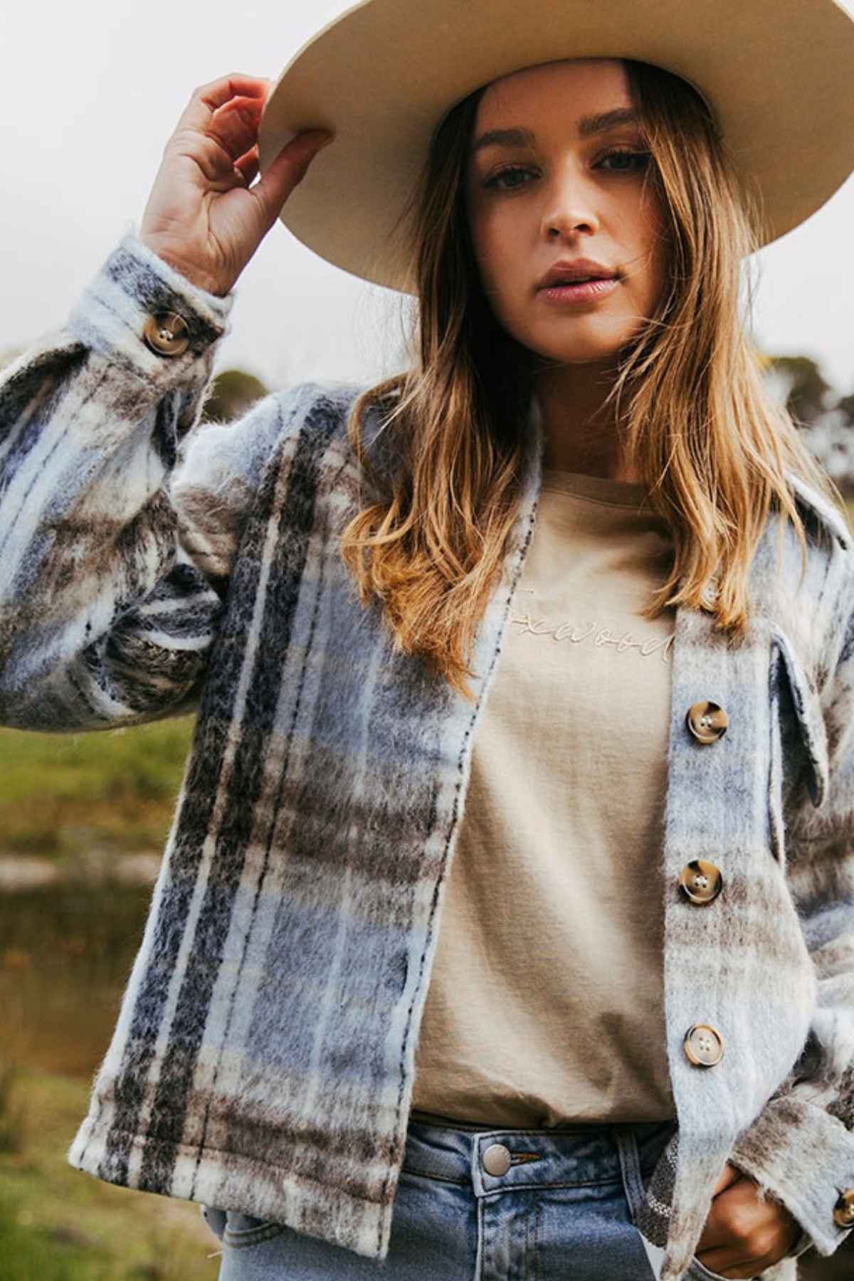 A woman stands outdoors in a Foxwood Rhodes Check Coat, touching her wide-brimmed hat and looking at the camera.