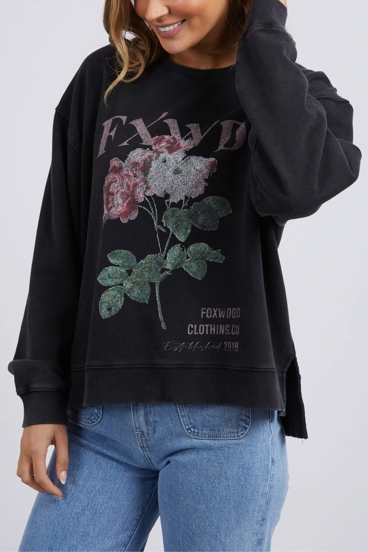 Wearing the Foxwood Rosa Crew—black with a floral chest print and "FOXWOOD CLOTHING CO" text—a person stands against a plain background, paired with blue jeans and a slight smile.