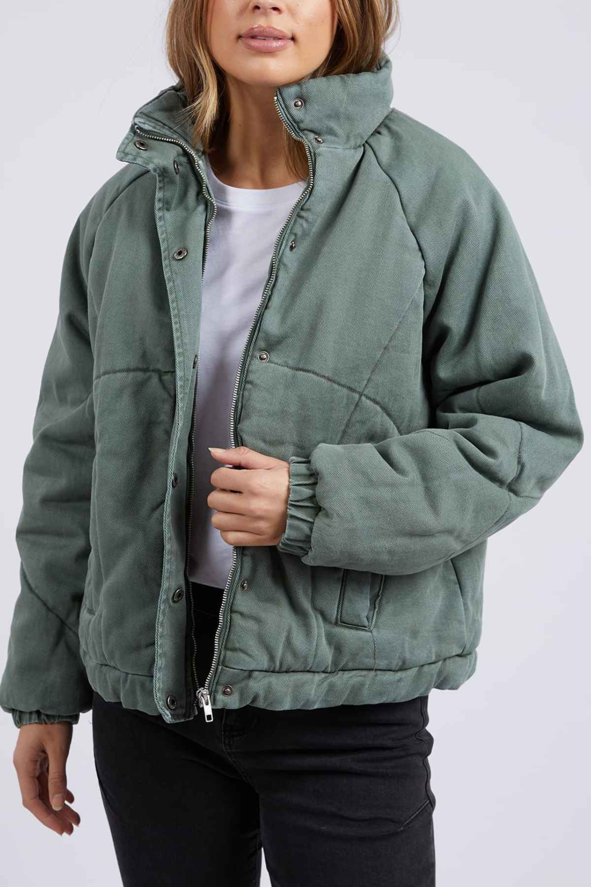 A person wearing the Foxwood Rosalee Denim Jacket, a cozy green cropped slouchy cotton denim, partially unzipped over a white t-shirt and black pants, stands against a plain light background holding one side of the jacket.