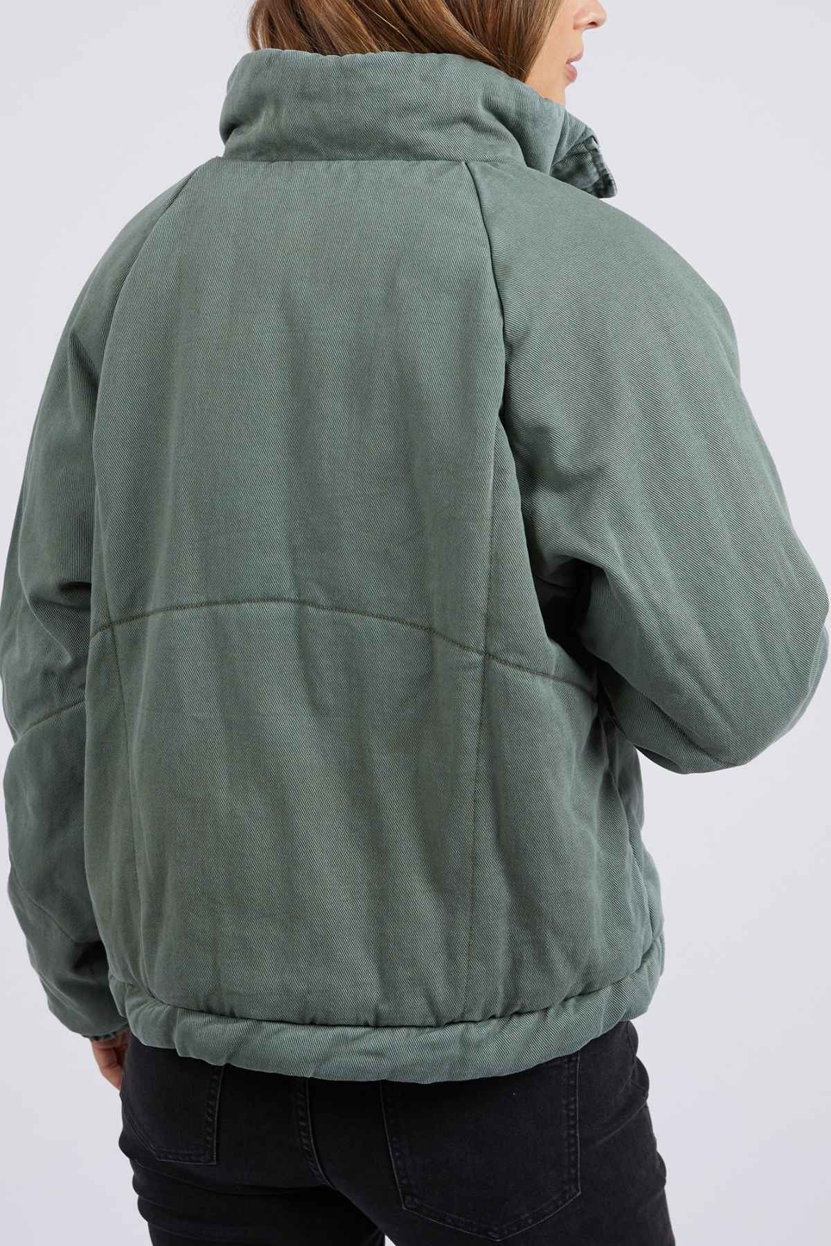 A person is shown from the back wearing the Foxwood Rosalee Denim Jacket in sage green with a cozy cotton denim feel. The high collar pairs well with black jeans, and their brown hair is visible while their face is turned away.