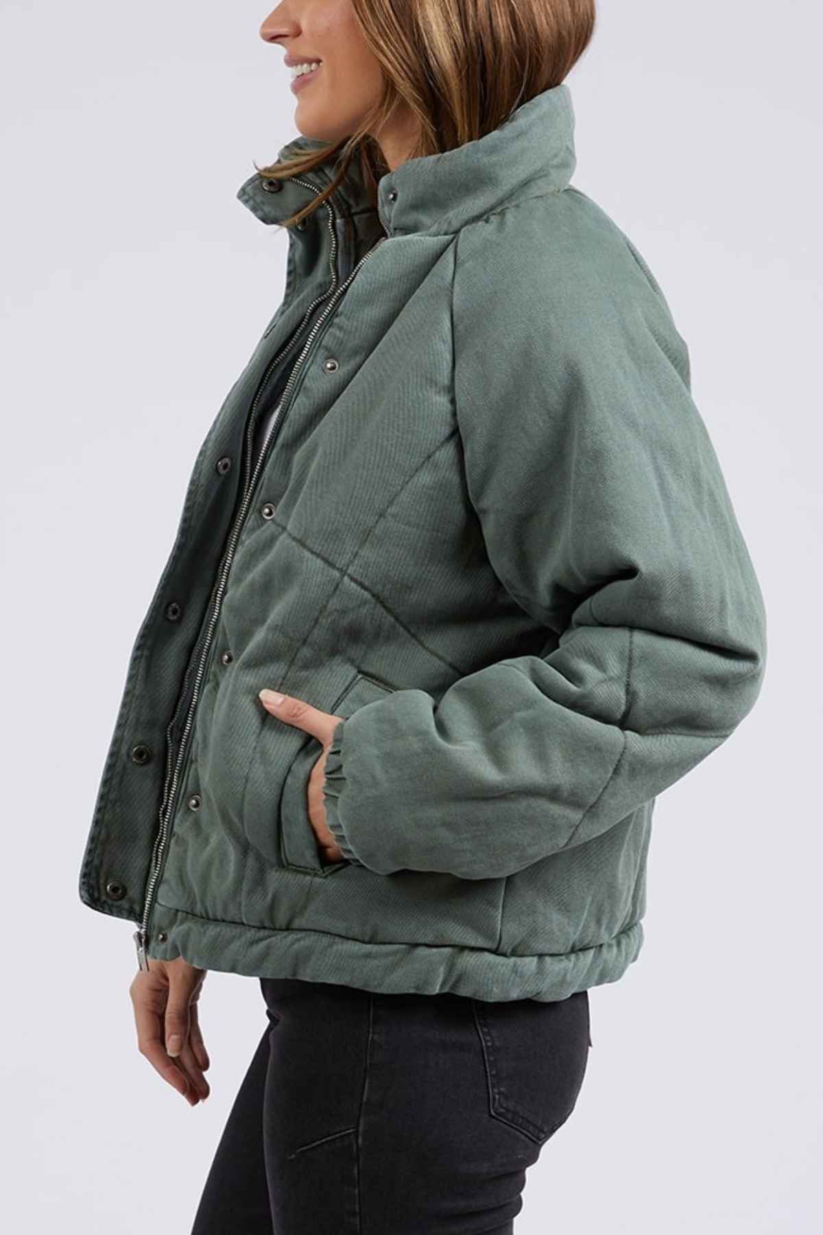 Woman in profile, smiling, wears the Foxwood Rosalee Denim Jacket—a sage green quilted puffer with snap buttons and side pockets—paired with dark skinny jeans. Background is plain and light-colored.