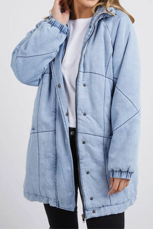 A woman wears the Foxwood Rosalee Longline Jacket in light blue over a white t-shirt and black pants. The padded, 100% cotton denim jacket features a front zipper, snap buttons, and elastic cuffs. Her face is partly out of frame.