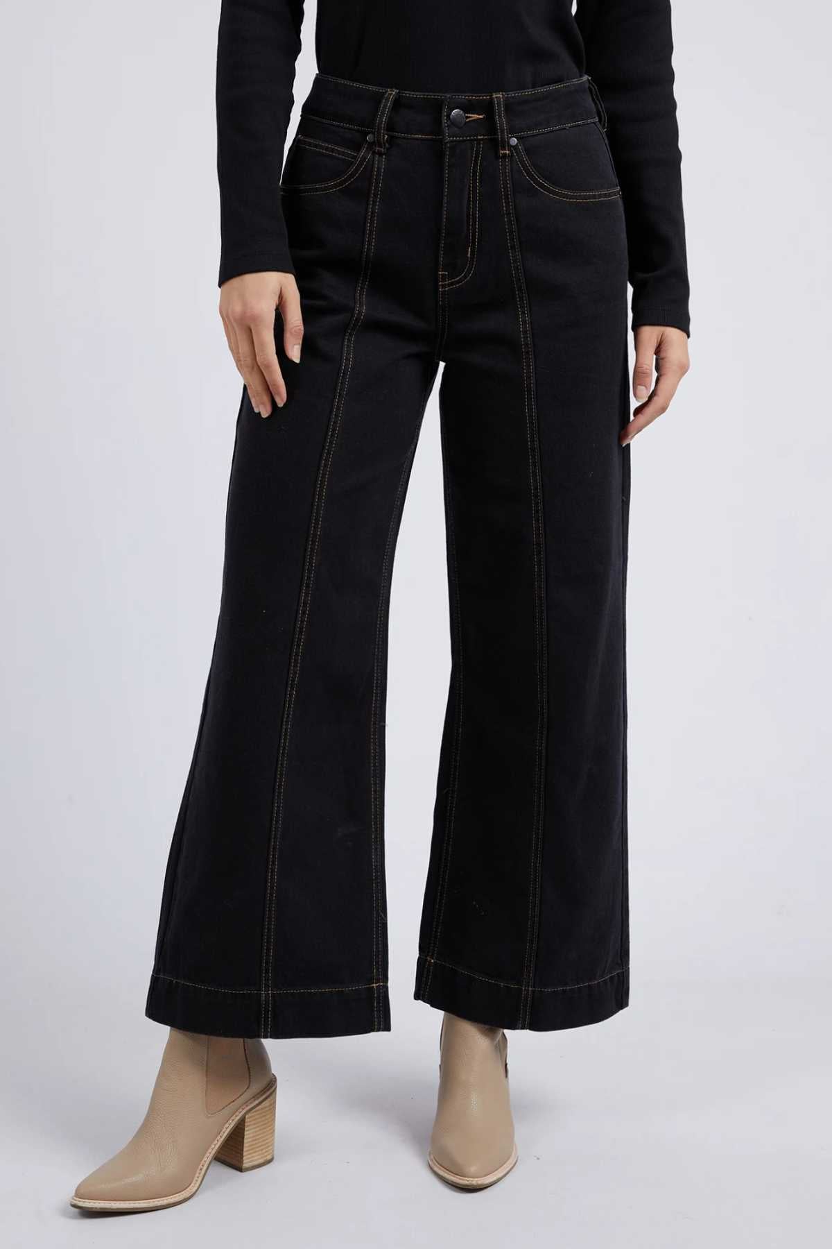 Someone in Foxwood Royal Wide Leg Jeans with contrast stitching and a black top stands against a plain light gray background, finishing the look with tan ankle boots featuring a block heel.