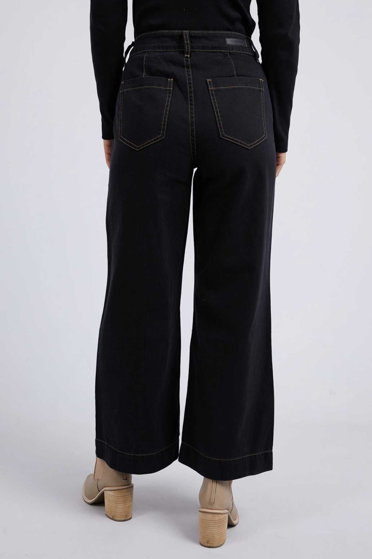 A person is seen from the rear wearing Foxwood Royal Wide Leg Jeans, characterized by high-waisted, wide-leg black denim with contrast stitching. They complement the look with beige heeled ankle boots and a black top against a plain light gray background.