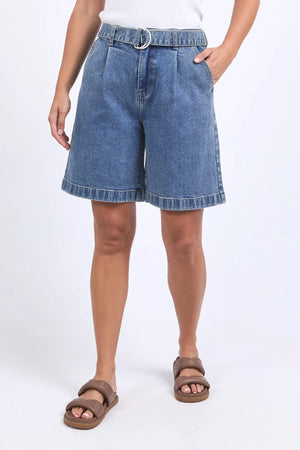 Wearing a white top, Foxwood Saint Short denim shorts with a belt, and brown sandals, a person stands with one hand in their pocket against a plain light background.
