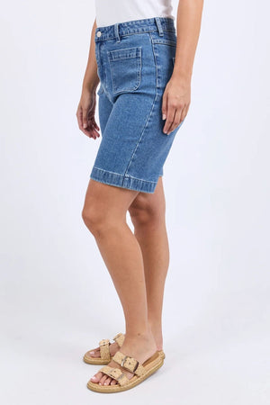 Someone stands against a plain light background, dressed in a white top, the knee-length blue denim Foxwood Sarah Short, and beige cork-sole sandals.