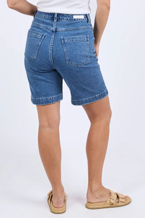 A person faces away, wearing the Foxwood Sarah Short knee-length denim shorts, a white top, and beige woven sandals against a plain light background.