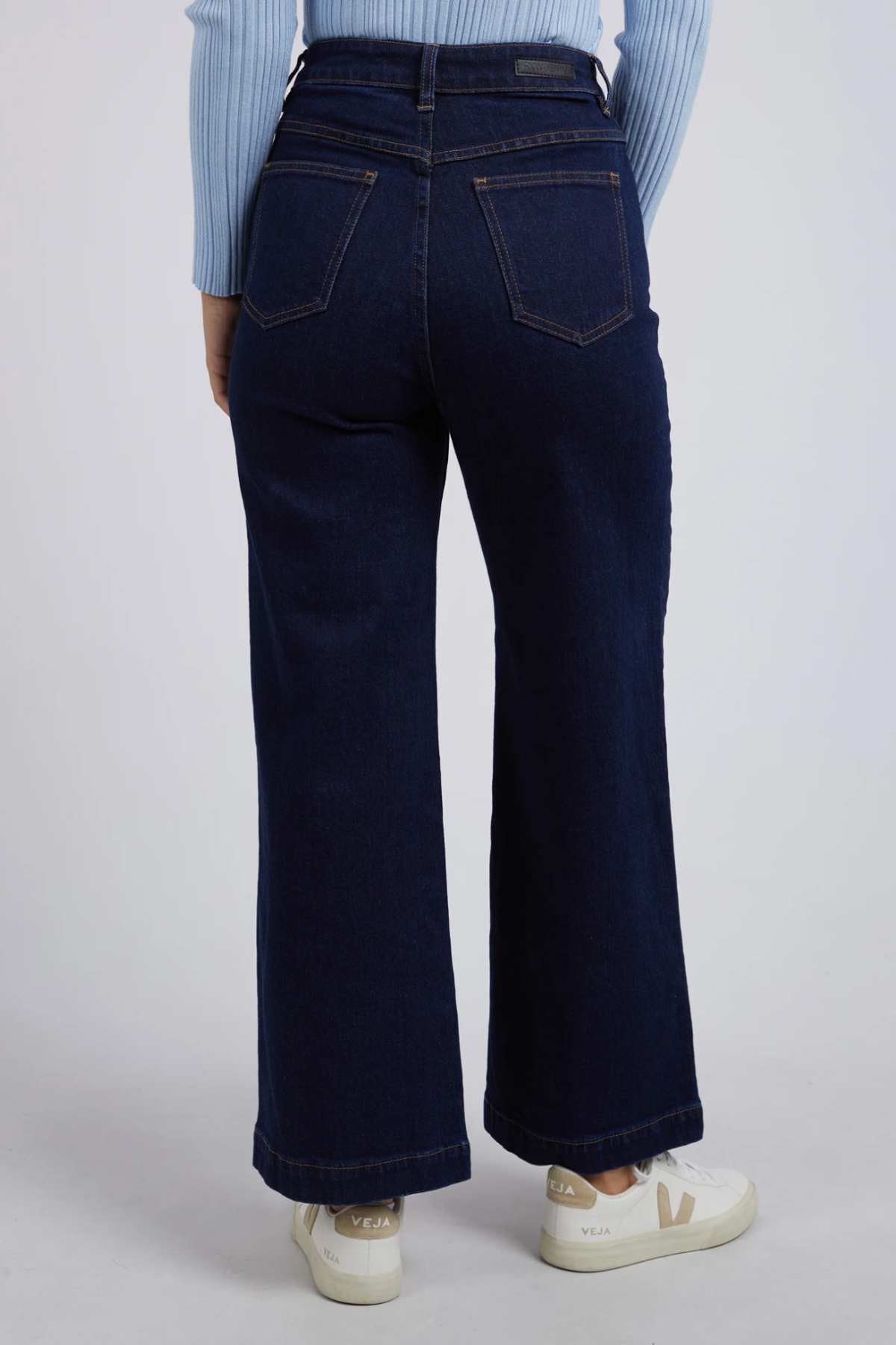 A person wearing Foxwood Sarah Wide Leg Jeans in dark blue with a high-rise waist and a light blue ribbed sweater. They are standing facing away from the camera, showcasing the back of the cotton elastane jeans and pockets. The person is also wearing white sneakers with visible branding.