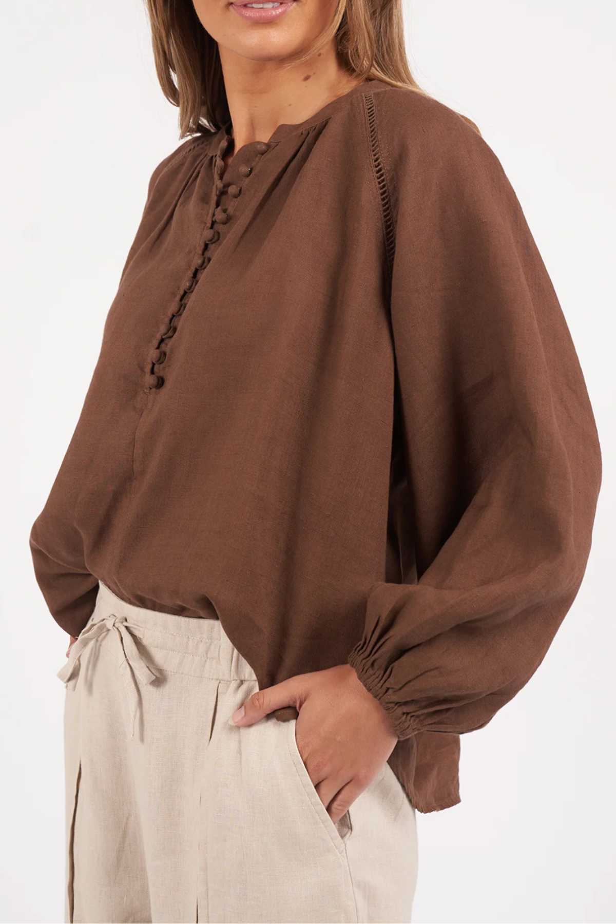 A person models the Foxwood Sardinia Blouse, a long-sleeved, loose brown top with fabric-covered buttons and elastic balloon sleeves, paired with high-waisted light beige drawstring pants against a plain white background.