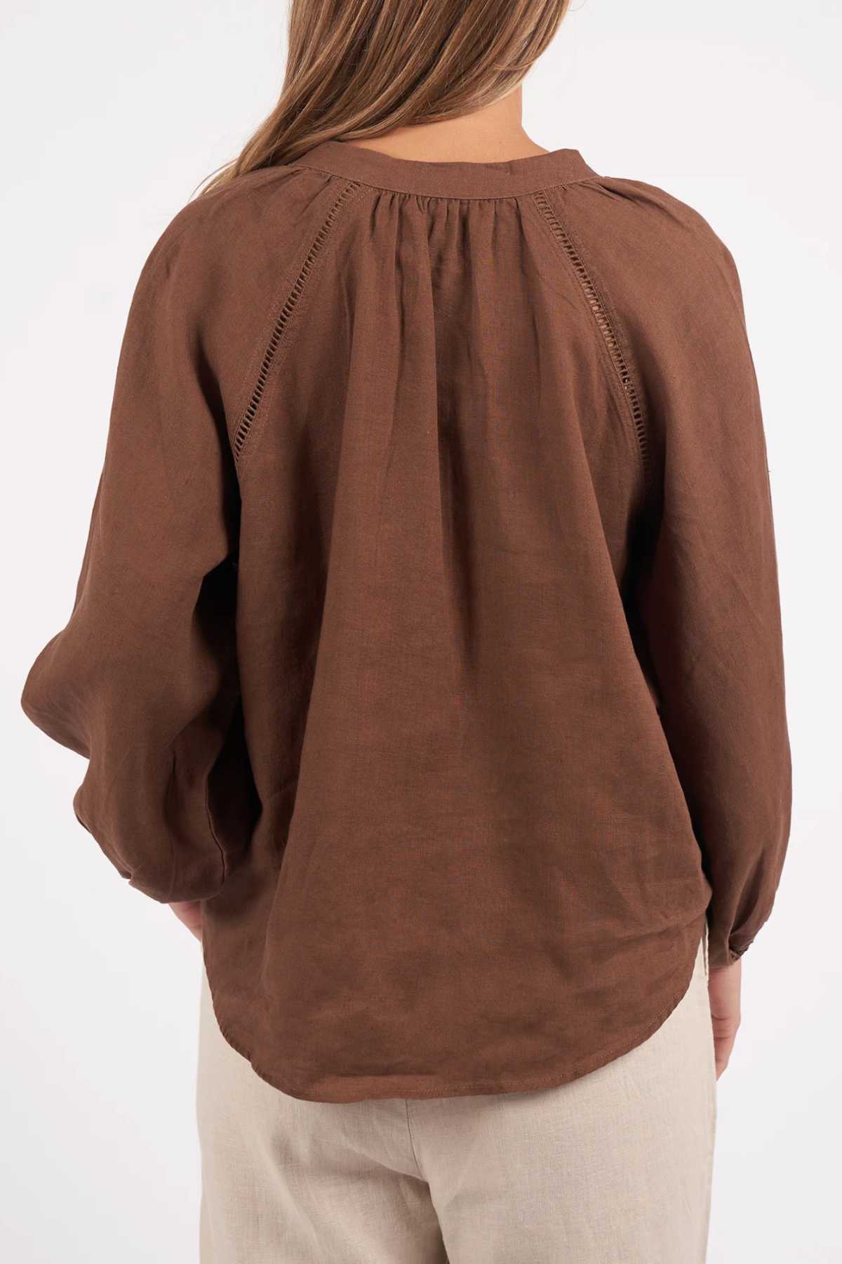 A woman stands with her back to the camera, highlighting the Foxwood Sardinia Blouse—a loose, long-sleeved brown top with gathered details and fabric-covered buttons—paired with light pants. Focus is on her upper body and the blouse's back.