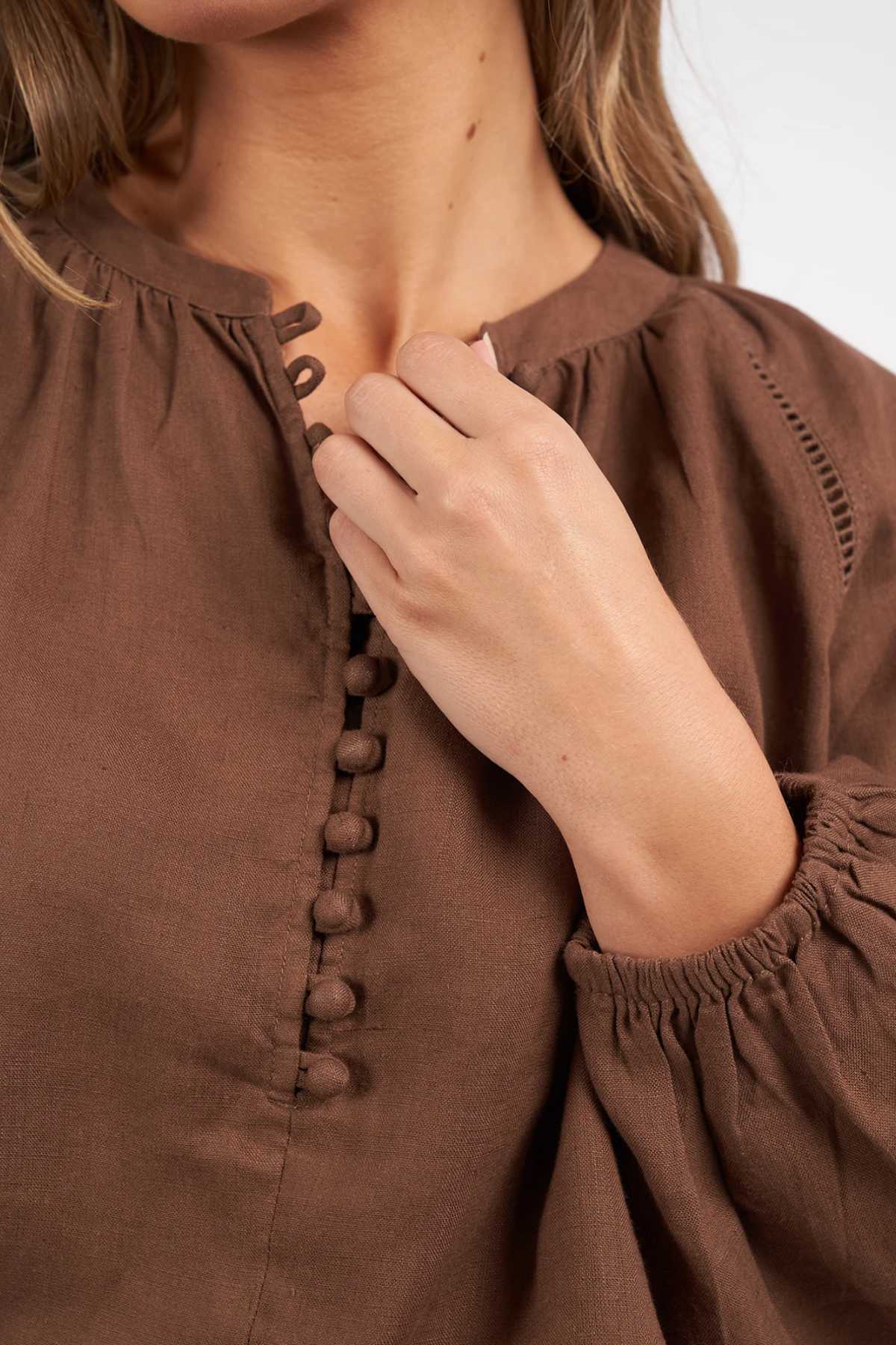 A woman wears the Foxwood Sardinia Blouse, detailed with fabric-covered buttons down the front. Only her lower face, neck, and upper body are visible as she softly touches her neckline.
