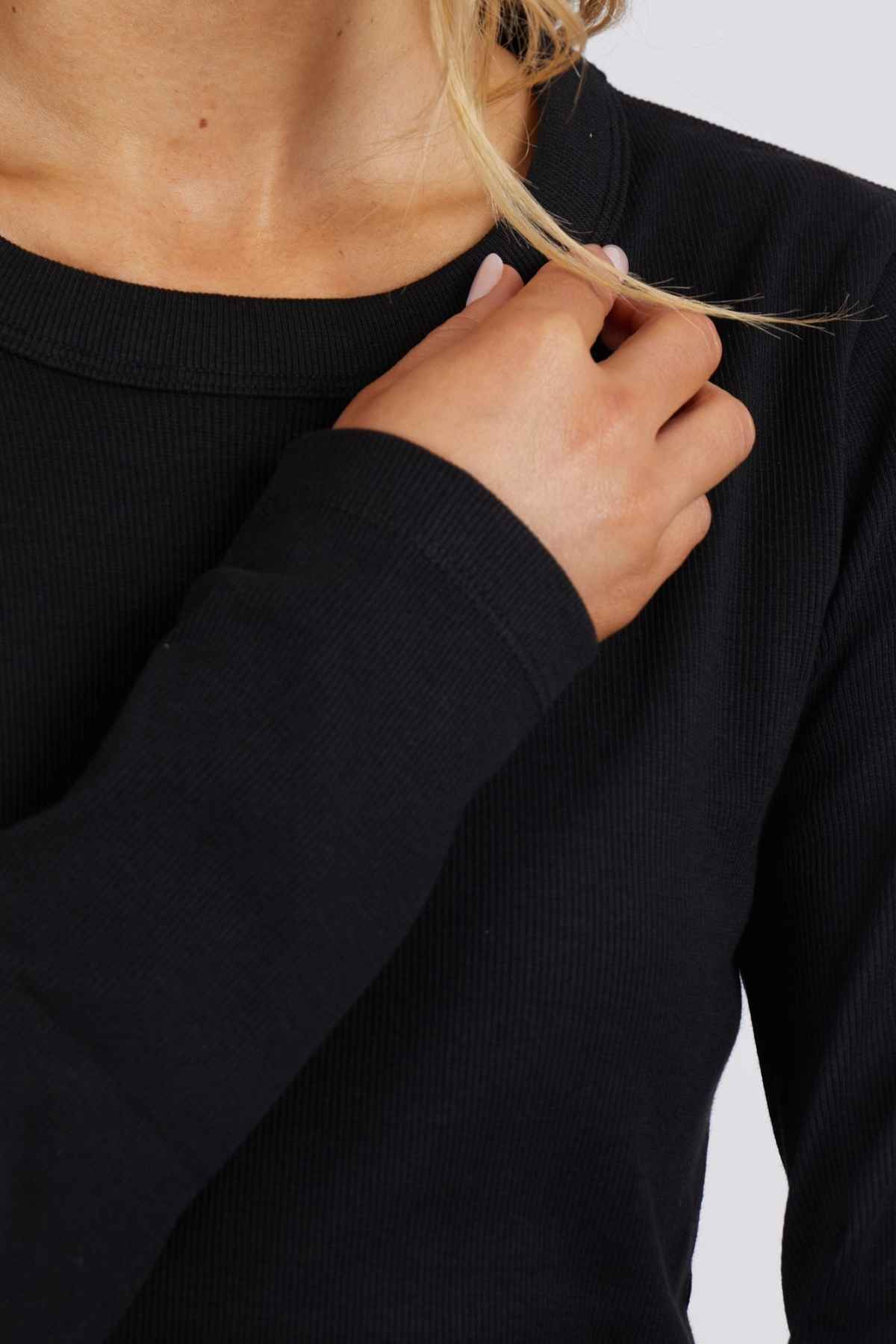 A person wearing a black Foxwood Scoop Long Sleeve Rib sweater holds a strand of their light blonde hair, with the image focusing on the upper torso and hand near the shoulder against a light gray background.