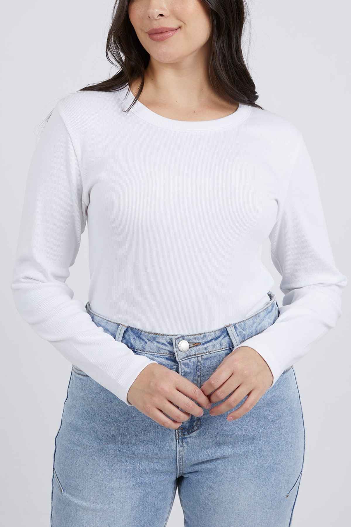 A person with long dark hair, wearing a plain white Foxwood Scoop Long Sleeve Rib shirt tucked into light blue high-waisted jeans, is standing against a plain background. The Foxwood scoop neckline adds charm as they smile gently, hands resting at their waist.